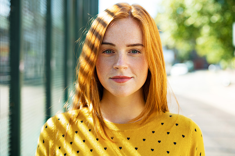 A young woman with striking red hair stands near a fence, her expression reflecting contemplation and poise.
