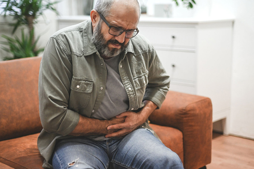 A man seated on a couch, appearing uncomfortable, with his hand on his abdomen, indicating he is in pain