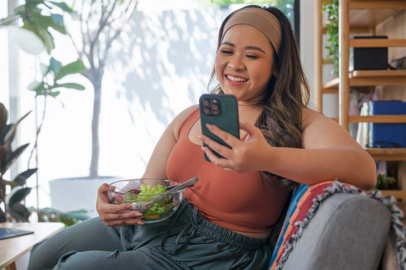 A woman holding a salad and looking at her phone, browsing Labcorp OnDemand’s Personal Wellness Program.