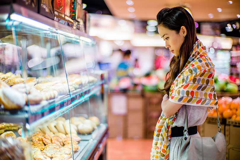 A woman in a grocery store looking at a display case of food, unsure if her digestive symptoms are related to celiac disease or IBS and whether she should be eating gluten-free food.