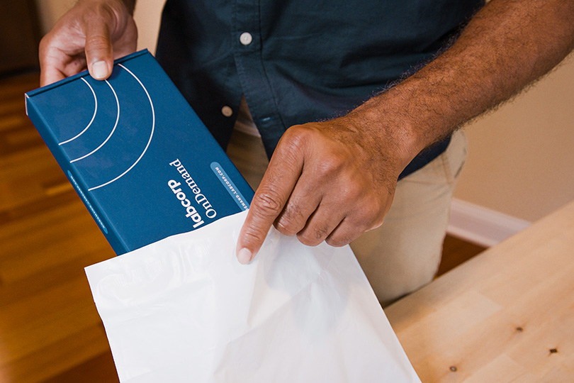 A man displays a white bag with a blue box home collection kit, illustrating the importance of home collection testing for colon cancer screening.