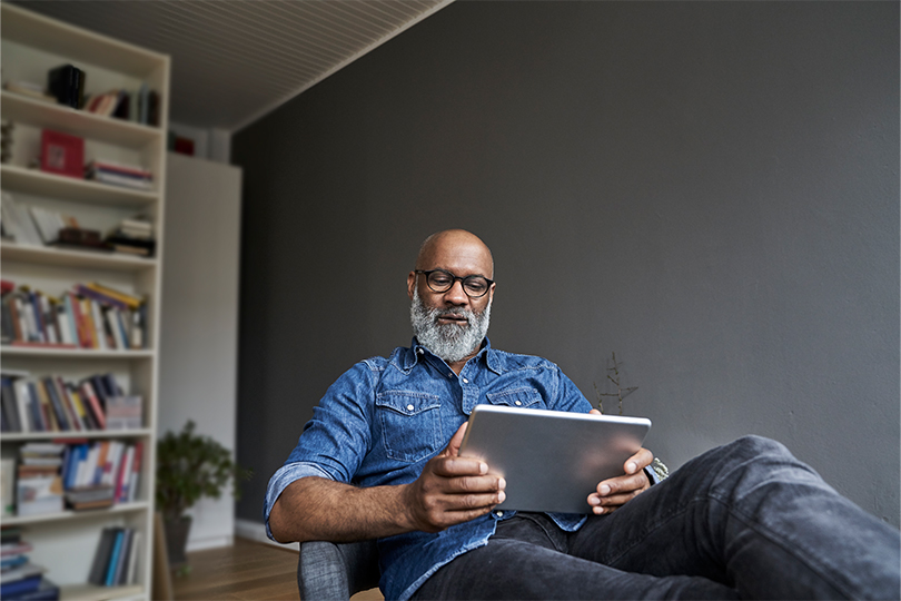 An older Black man wearing glasses sitting in a chair inside an office holding a tablet, reading about low testosterone