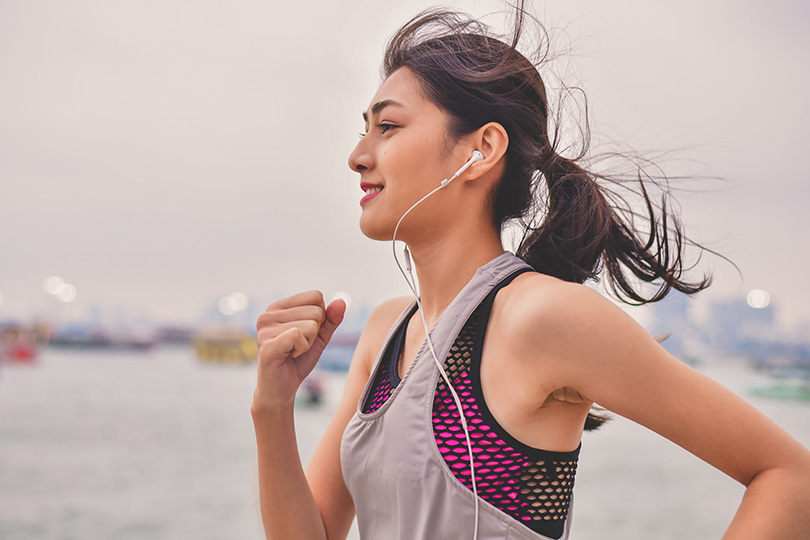 A woman wearing earbuds runs near the water, illustrating the benefits of exercise for increasing good cholesterol.