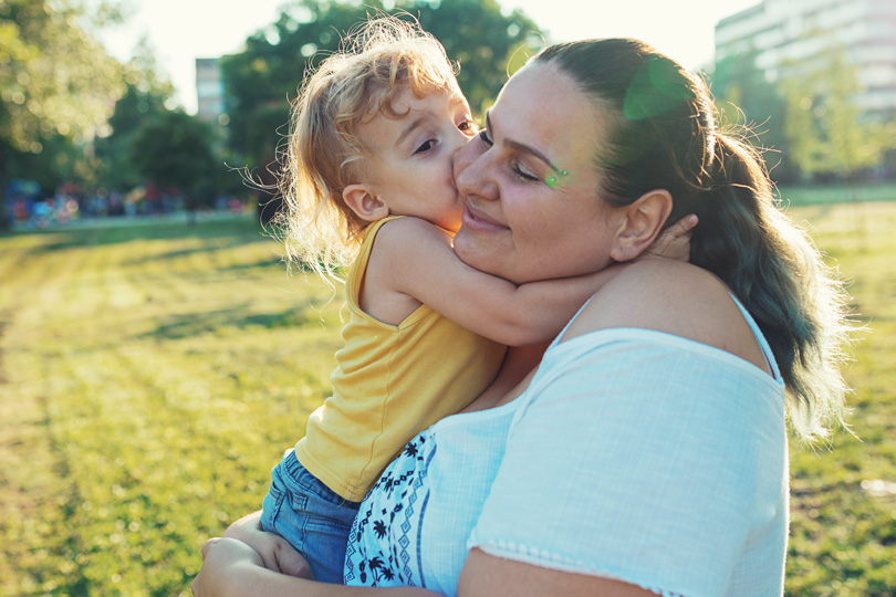 A woman standing outside holding a child, smiling and hugging.
