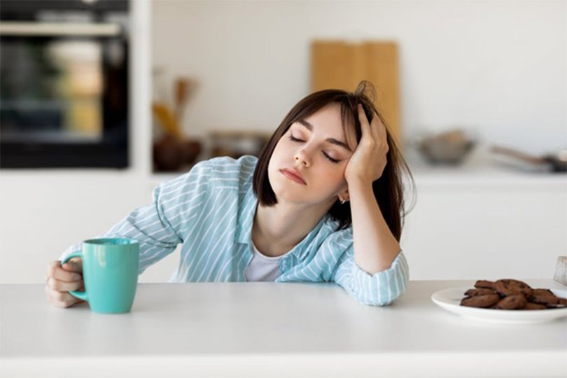 A woman in her kitchen closing her eyes and resting her head in her hand, looking fatigued, a possible symptom of abnormal MCV levels.
