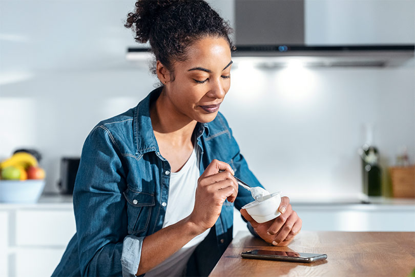 A woman sits at a table reading about what prediabetes is while eating yogurt.