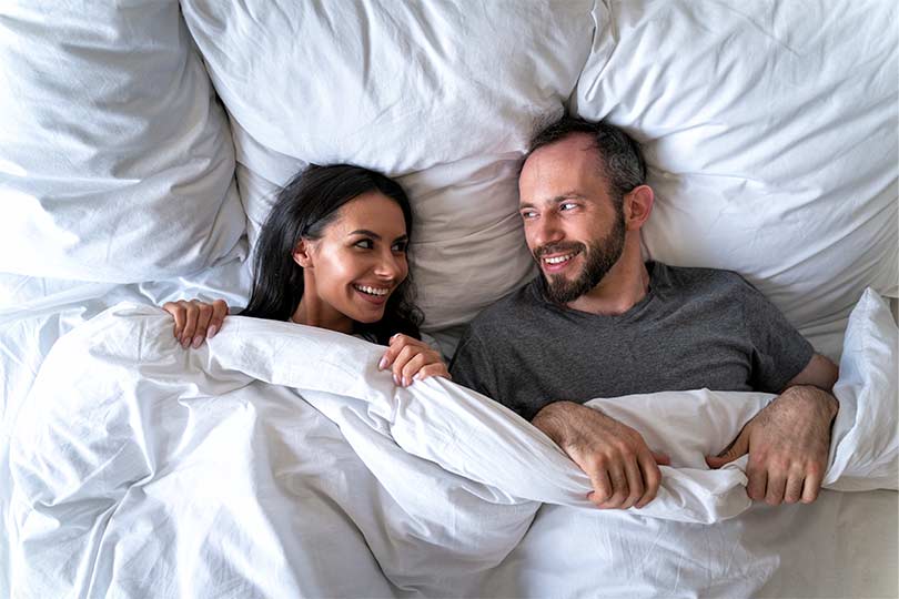 A man and a woman laying in bed together smiling at one another, engaging in open communication about herpes and STI testing.