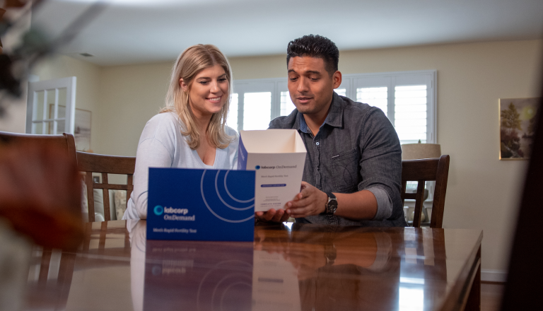 Man and woman at table examining Labcorp OnDemand at-home men's rapid fertility test box, showing interest and engagement