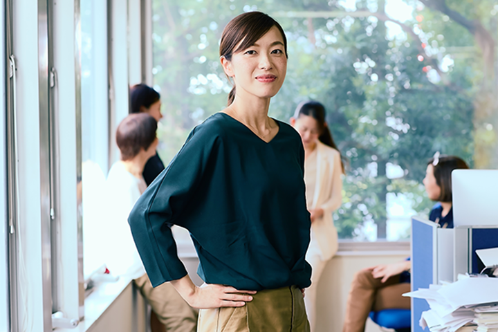 Woman standing with hand on hip in office, other women in background, windows and papers visible.
