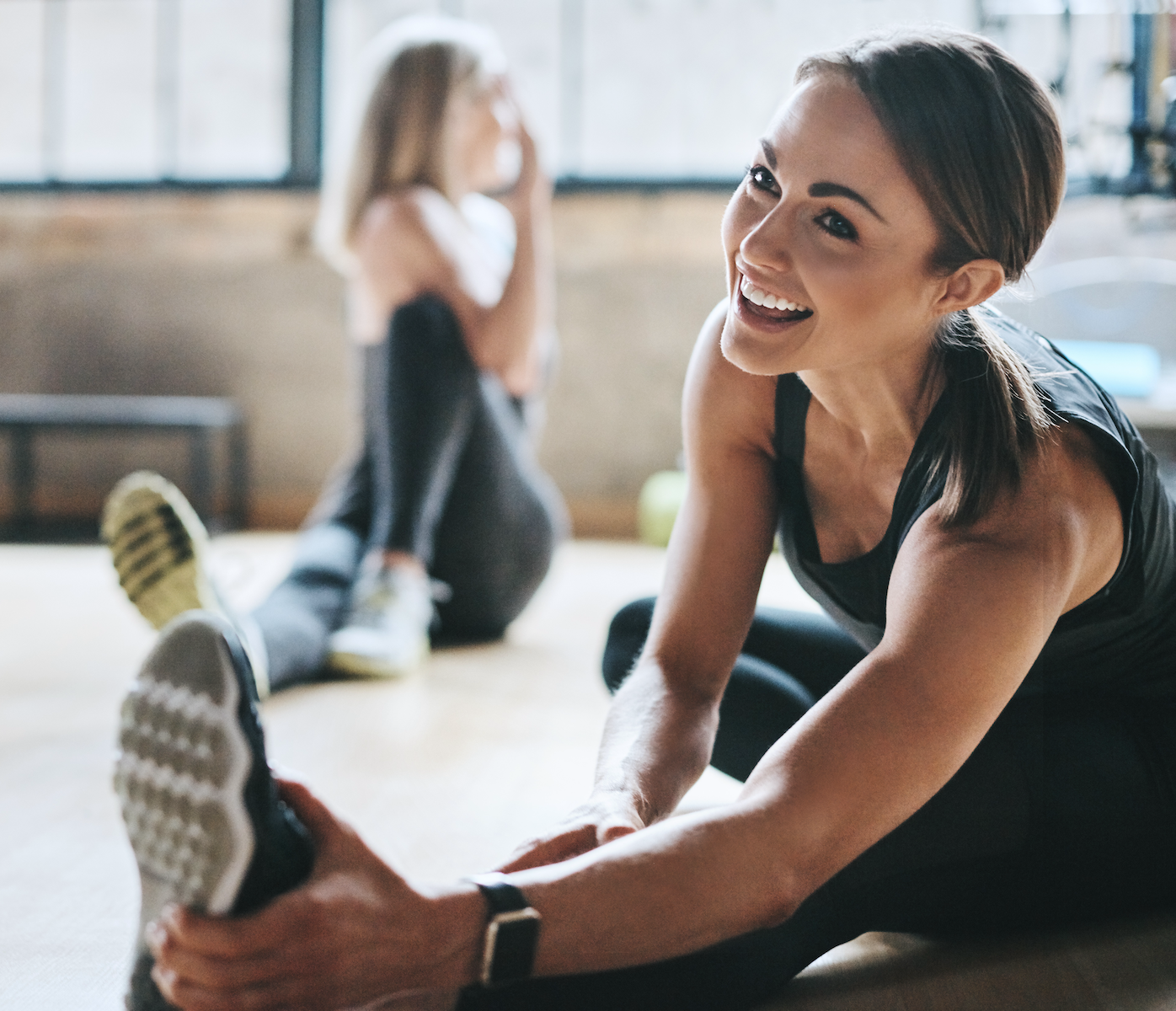 Woman stretching in fitness center, wearing fitness watch and sneakers. Another woman stretching in background.