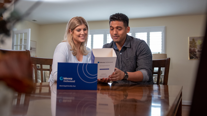 Man and woman at table examining Labcorp OnDemand at-home men's rapid fertility test box, showing interest and engagement
