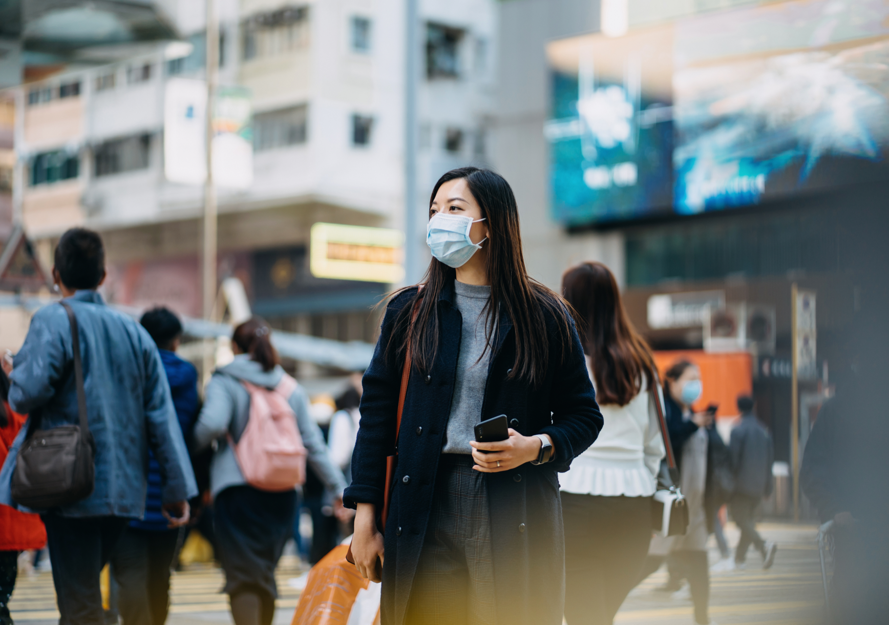 Masked woman holding phone in busy city street with pedestrians walking around her.