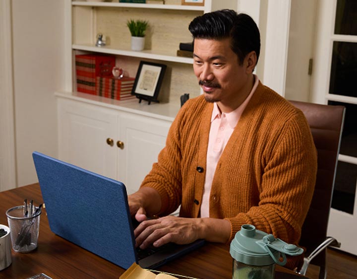  A middle aged man in an orange sweater working on his laptop in a home office.