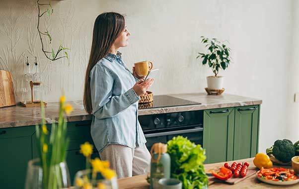 Woman standing in a kitchen holding a cup and a smartphone, appearing relaxed while researching custom pregnancy testing.