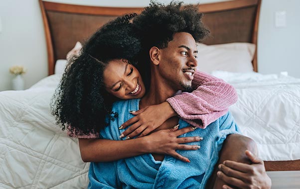 A couple sits on the floor by a bed, embracing and smiling warmly.