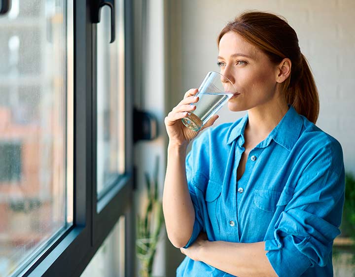 A woman drinks water while looking out the window in her home.