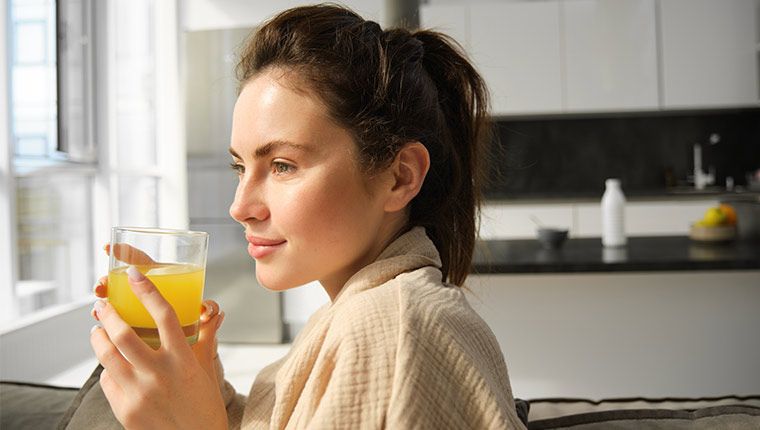 A woman sitting and holding a glass of orange juice looking out the window.