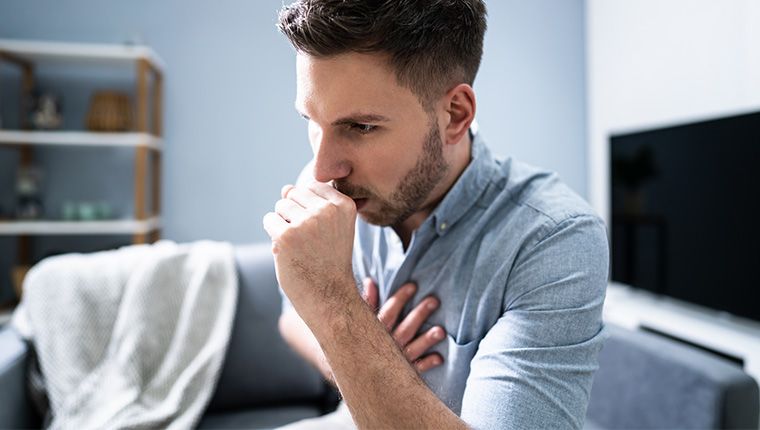 Young man sitting on couch in living room, feeling sick and coughing.